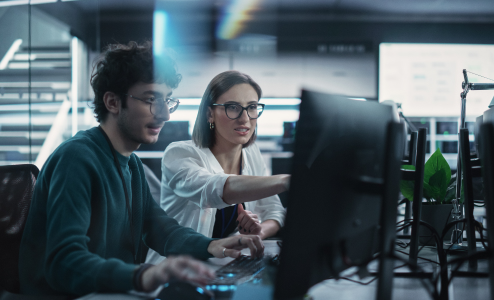 Two young professionals, a man and a woman, collaborate at a desk, looking at computer monitors with production data. The room has a modern tech aesthetic.